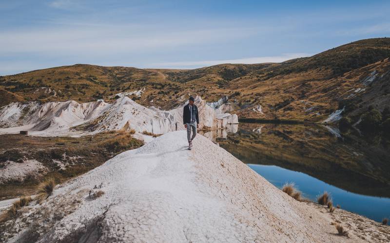 man walking near blue lake walk new zealand surrounded by mountains min