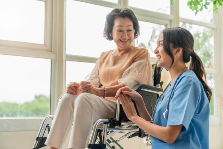 asian young caregiver caring her elderly patient senior daycare handicap patient wheelchair hospital talking friendly nurse looking cheerful nurse wheeling senior patient 768x513 1