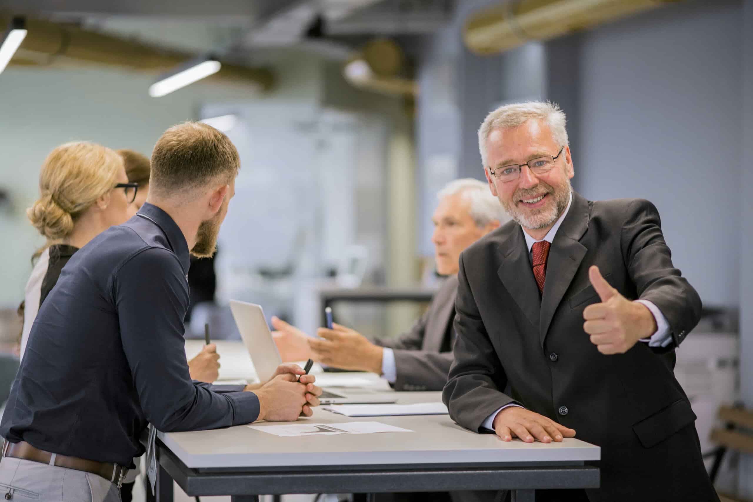 smiling senior businessman showing thumb up sign front businesspeople discussing office scaled